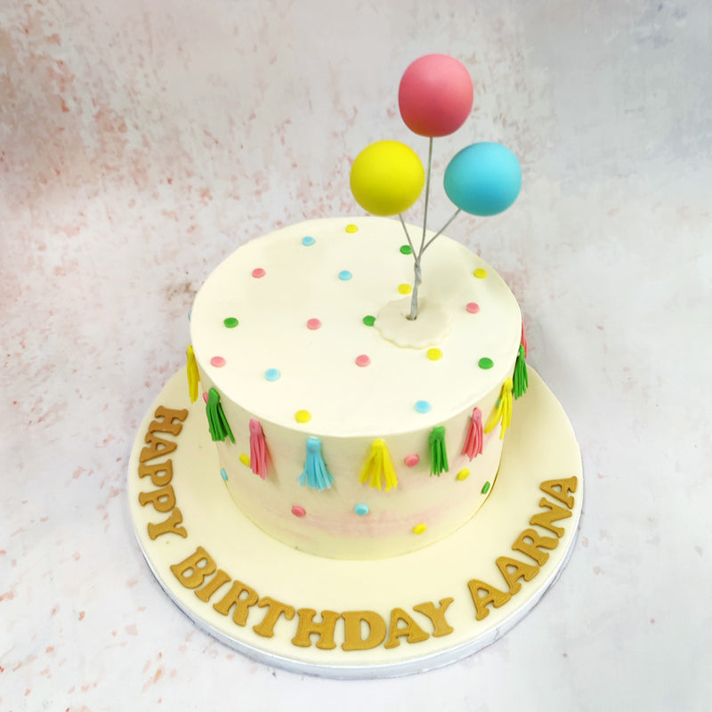 The surface of this Baby Shower Cake is a canvas of soft, creamy hues, punctuated by vibrant pops of color in the form of hand-crafted tassels and balloon-like spheres. 