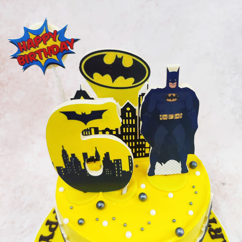 The centrepiece of this Batman Comic Theme Cake is a towering Batman silhouette, standing tall and proud against the backdrop of the iconic Bat-Signal. 