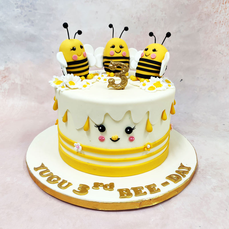 The front of this Birthday Cake For Kids features a sweet little face with blushing cheeks, fluttery lashes, and a tiny beak, turning the entire cake into a character of its own. Yellow stripes and piped floral accents wrap around the base, while teardrop-shaped honey drips fall gently from the top edge—adding texture and that sticky-sweet honeycomb vibe without overwhelming the design.
