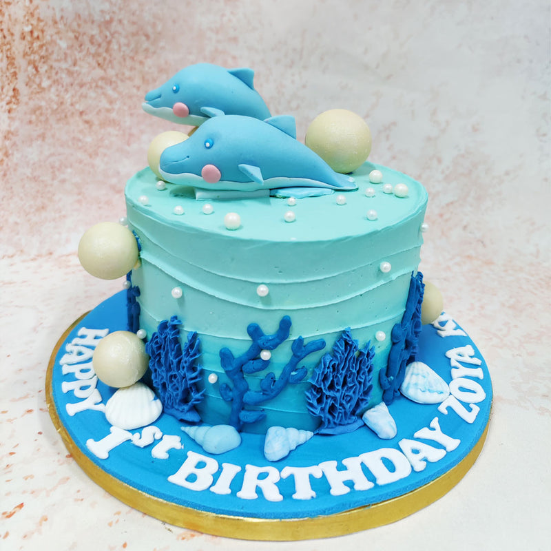 Playfully perched atop this Birthday Cake For Kids are two adorable blue dolphins with charming pink cheeks and friendly expressions, seemingly caught mid-leap as they frolic through imaginary waves. 
