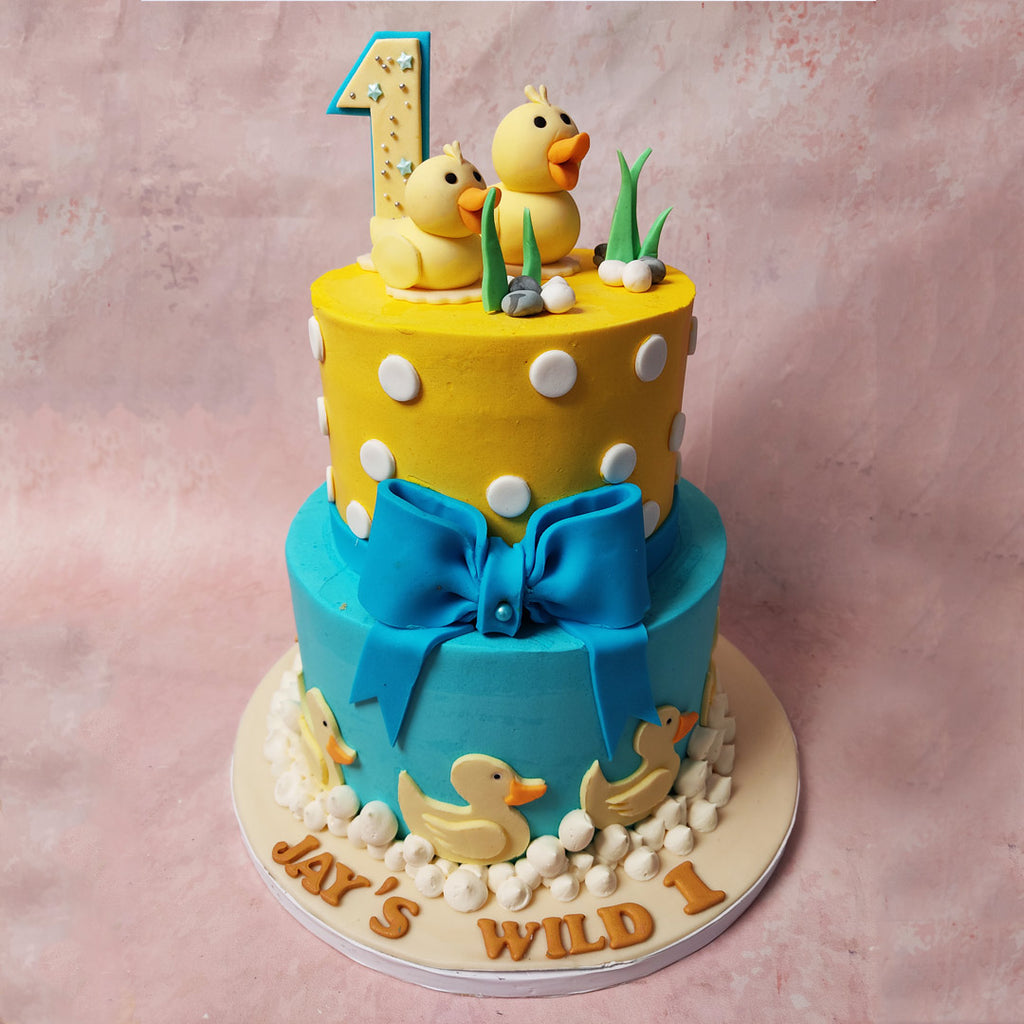 The lower tier of this Duck Theme Cake boasts a vibrant blue base, adorned with ducks waddling along the waves of a stream, crafted from stiff white buttercream peaks. 