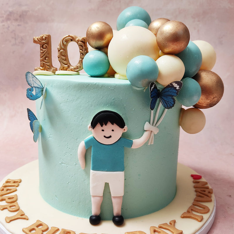 This Little Boy and Balloons Cake features a soothing blue hue, with a young lad at the centre holding onto a collection of chocolate balloons in white, gold, and blue. 