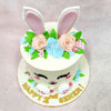 The front of this Bunny Face Cake is brought to life with bright, beaming eyes, rosy cheeks, and a cheerful smile—topped off with tall ears peeking through a floral crown of soft peach roses, blue blossoms, and hand-piped leaves.