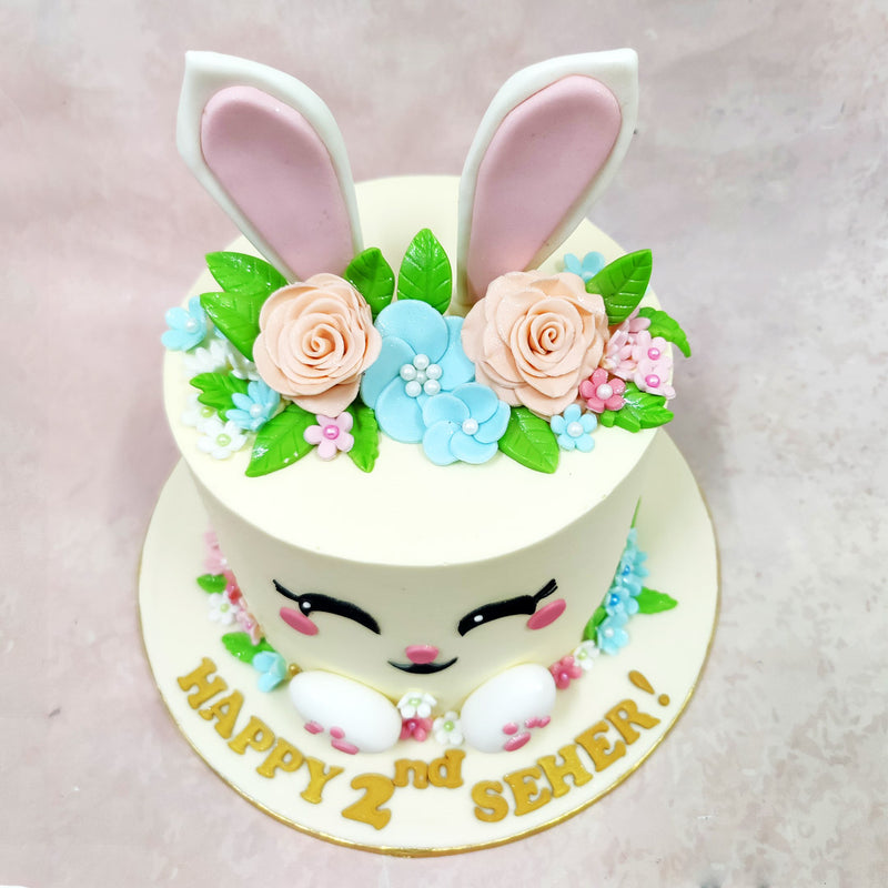 The front of this Bunny Face Cake is brought to life with bright, beaming eyes, rosy cheeks, and a cheerful smile—topped off with tall ears peeking through a floral crown of soft peach roses, blue blossoms, and hand-piped leaves.