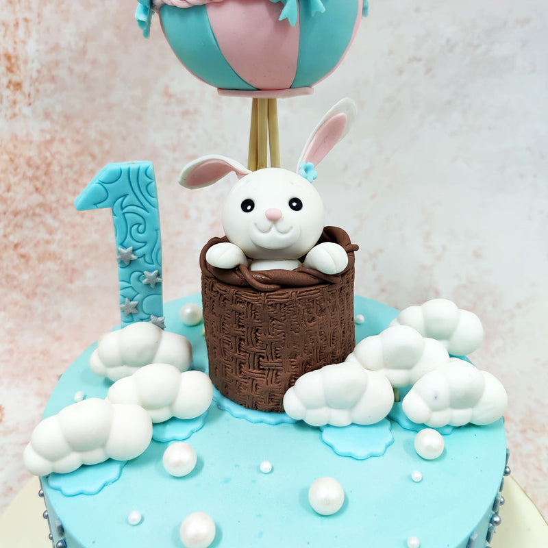 The centrepiece of this Bunny Hot Air Balloon Cake features an adorable white bunny peeking out from a textured chocolate basket, beneath a charming pink and turquoise striped balloon adorned with delicate bows and braided details.