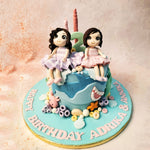 This Cake For Twin Sisters features two adorable girls sitting atop, each dressed in charming, striped dresses, capturing the innocence and joy of childhood. 