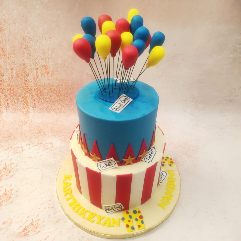 The top tier of this Circus Theme Cake features a bright blue base, adorned with playful details like tickets and golden stars, evoking the nostalgic feel of a big-top circus. 