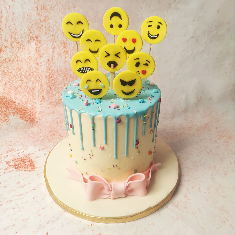 The  Emoticon Cake is adorned with a smooth, light blue drip icing cascading down the sides, adding a dynamic and visually appealing element. 