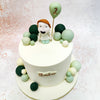 The centrepiece of this Green Balloon Theme Cake is an adorable figure of a smiling child, holding a pastel green balloon that adds a playful touch to the overall composition. 