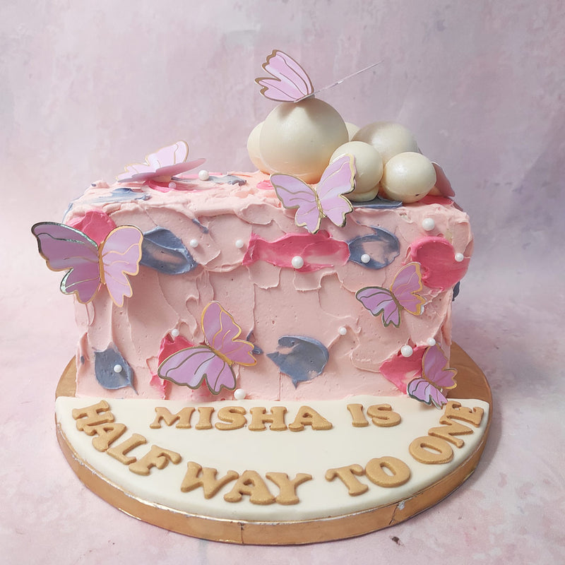 The light pink textured base of this Butterfly Half Birthday Cake represents the delicate nature of infancy, while the pink and blue buttercream smudges add a touch of playfulness and joy. 