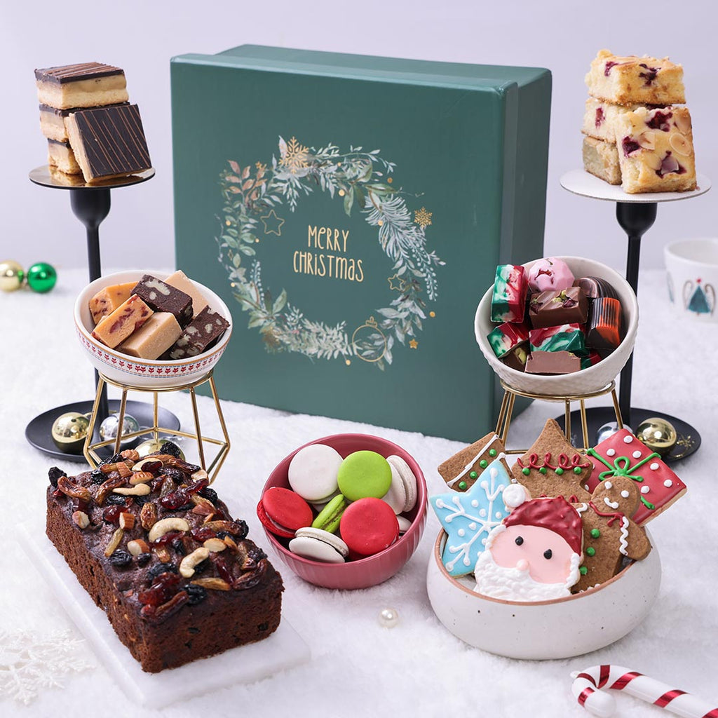 Assorted Christmas treats including cookies, macarons, and a cake in front of a 'Merry Christmas' gift box.