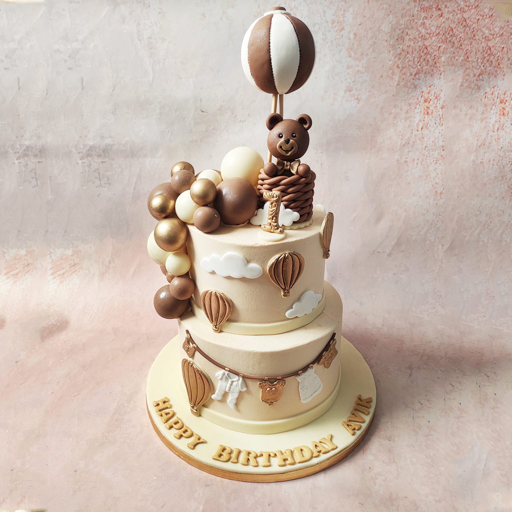 Graceful copper hot air balloons ascend through the sugary sky on this Hot Air Balloon Teddy Bear Cake, each carrying the hopes and dreams of a young heart.  Descend to the bottom tier of this Teddy Balloon Cake, where a bronze clothesline cradles baby clothes in hues of white and copper. 