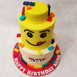 A two tier Lego cake design, with the bottom tier in a neutral shade, embellished with edible Lego blocks bordering the bottom and strewn on top.