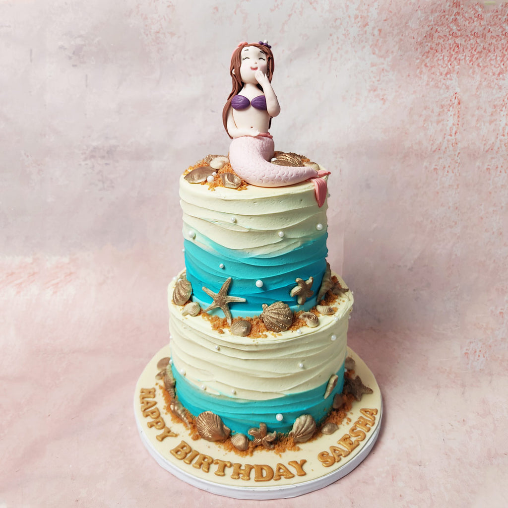 The buttercream canvas on this Two Tier Mermaid Theme Cake, shifting from a deep blue to a serene white in wavy patterns, mirrors the ebb and flow of the sea and the sandy shores. 