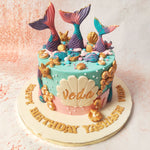 The vibrant tails of mermaids emerging from the surface of this Mermaid Seashell Cake are a captivating focal point, shimmering with iridescent hues of purple, pink, and blue, accented with golden tips that glisten like treasures of the deep. 