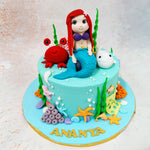 The centrepiece of this Mermaid and Shark Cake is a charming mermaid figurine, with flowing red hair and a shimmering teal tail, seated amidst a whimsical aquatic scene. 