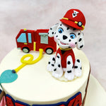 The round Mighty Pups Cake features adorable figurines of two popular Paw Patrol puppy characters—one sporting a red firefighter outfit with matching helmet and another in police attire sitting alongside. 