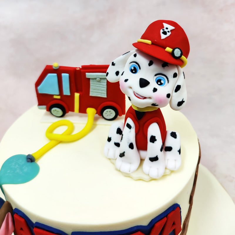 The round Mighty Pups Cake features adorable figurines of two popular Paw Patrol puppy characters—one sporting a red firefighter outfit with matching helmet and another in police attire sitting alongside. 
