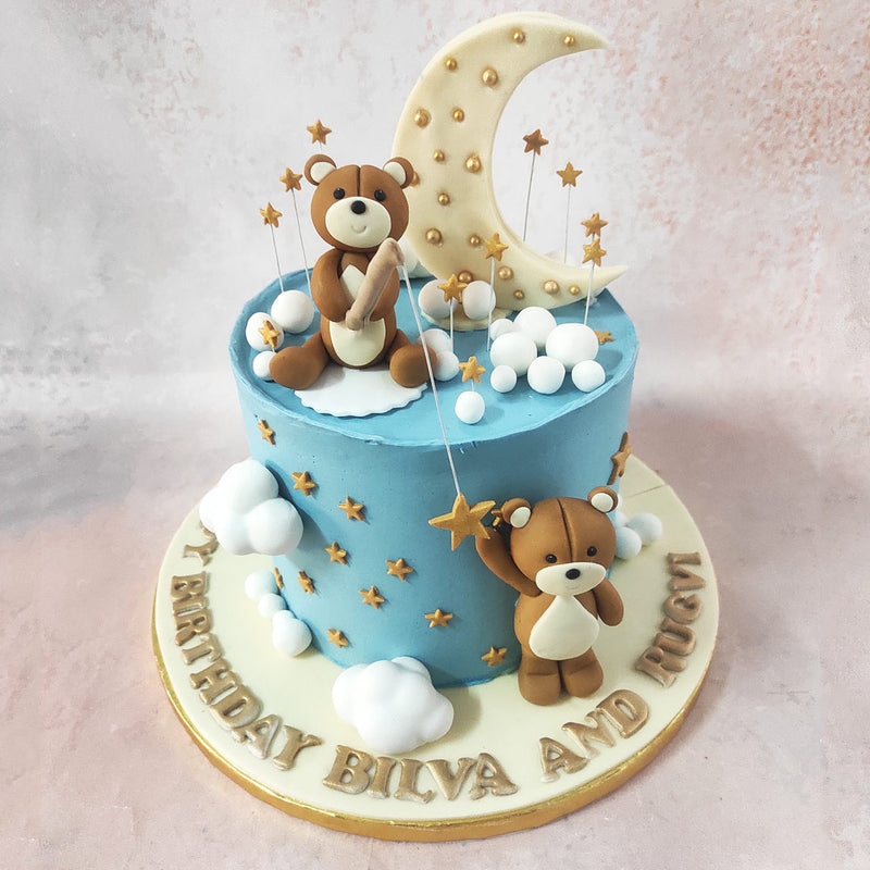 One teddy is perched on top of this sky theme cake, holding a fishing rod with a star-shaped lure dangling from the hook. The other teddy bear is below the teddy bear cake with stars, looking up at its friend with anticipation.