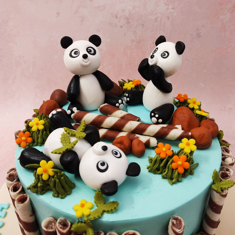 A cute panda cake complete with not one, not two, but three adorable, miniature figurines of pandas playing in a field of colourful flowers and rocks.