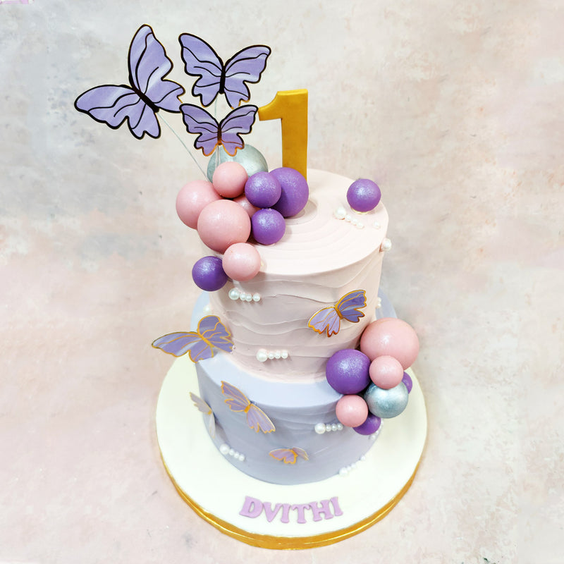 Two-tier birthday cake with purple and pink balloons, butterfly decorations, and the number '1' on a light background.