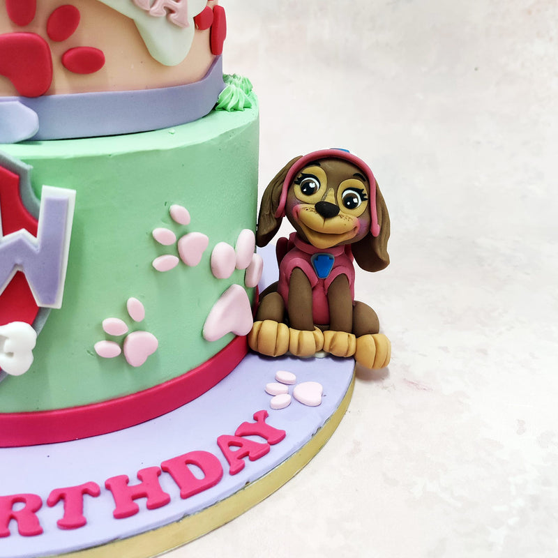 The bottom tier, featuring a vibrant mint green and pastel lavender base, showcases the iconic Paw Patrol badge front and centre, adorned with paw prints and a miniature dog collar detail, perfect for adding a touch of the series' canine charm to this Paw Patrol Zuma Cake. 