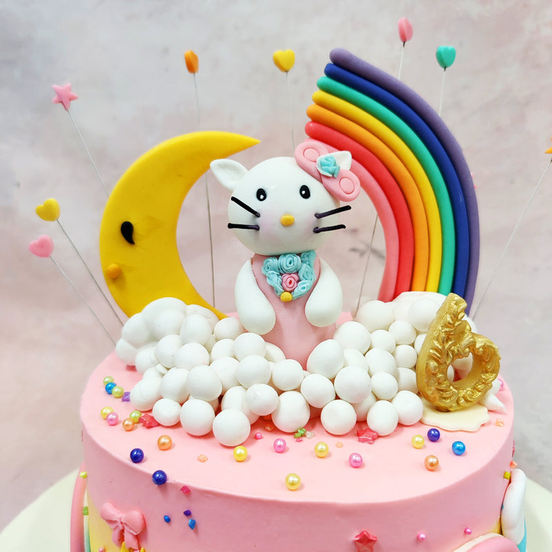 Floating atop a bed of whipped cloud puffs, the star of this Pink Kitty Cake sits sweetly with a tiny floral bow, pearl necklace, and the daintiest smile, surrounded by a golden crescent moon, a bold rainbow arc, and fluttering hearts on wires that seem to dance in mid-air.