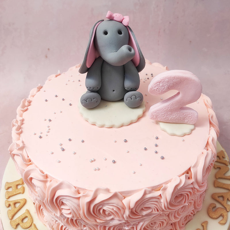 Perched atop this Pink Rosette Cake is the pièce de résistance—a meticulously sculpted baby elephant, its fondant form brought to life with artful shading and gentle curves. 
