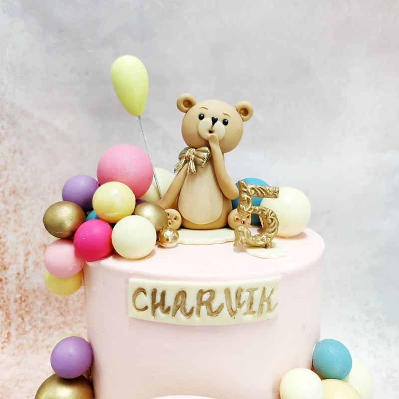 The centrepiece of this Pink Teddy Balloon Cake is an adorable fondant teddy bear, with a golden bow sitting proudly on top. 