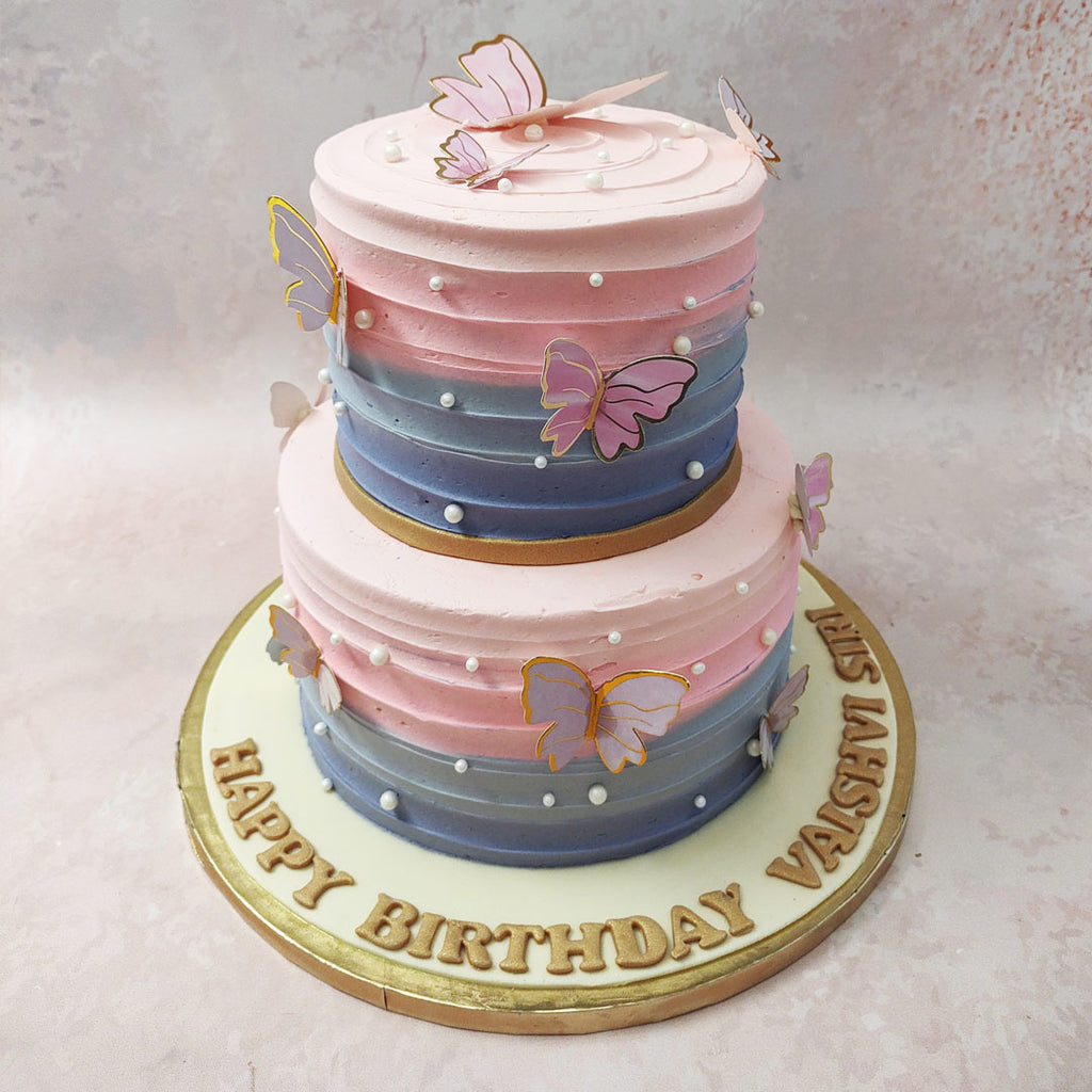 This enchanting two tier butterfly theme cake is adorned with delicate edible pearls, lifelike 3D butterflies, and a graceful gold ribbon that gracefully wraps around both tiers. 