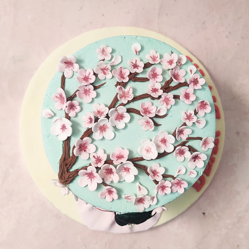 This flower tree cake features a three-dimensional figurine of a girl facing the sky blue base in a beautiful pink sundress as she reaches towards the realistic-looking edible Sakura tree blooming on this cherry blossoms cake creating the perfect imagery of spring.