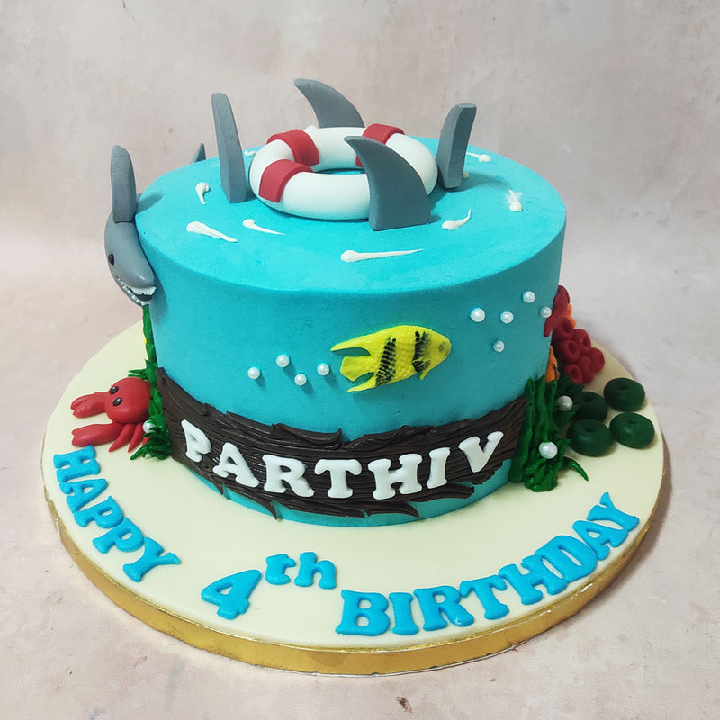 As you journey into the depths of this Shark Theme Cake, you'll encounter an edible wooden log at the centre of the blue base, proudly displaying the birthday child's name like a placard heralding their undersea kingdom. 