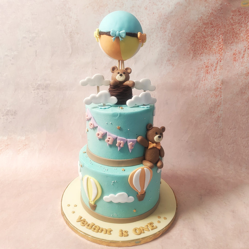 This two-tier Hot Air Balloon Teddy Bear Cake, bathed in the soothing hue of light sky blue, becomes a canvas of nostalgia adorned with fluffy white clouds. 