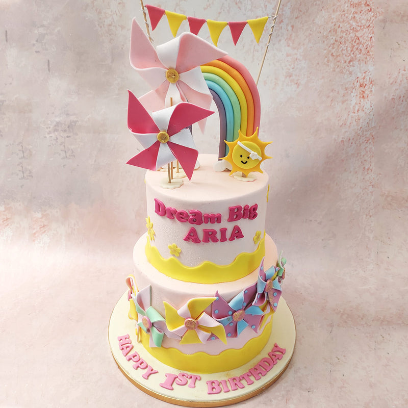 Around the bottom tier of this Pink Rainbow Cake, a merry parade of colourful pastel windmills twirled gracefully, adding a touch of whimsy to the scene.