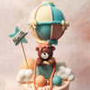 The bottom tier of this Two Tier Hot Air Balloon Teddy Bear Cake boasts a swirl of blue, peach, and cream buttercream piping, while the top tier features a smooth peach buttercream adorned with fluffy white clouds. 