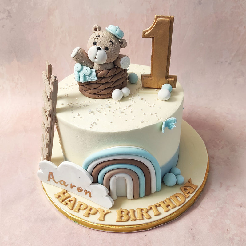 Blue icons of comfort and joy adorn this teddy bear in a basket cake,  from the blanket on the side of the basket to the ribbon at the base.