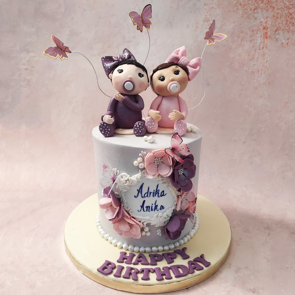 Set on a muted blue base, bearing resemblance to the comforting colours of our childhood rooms and embellished with pearls of buttercream at the bottom, this twins birthday cake for kids showcases an elegant wreath of flowers and butterflies at the centre near the name display. 