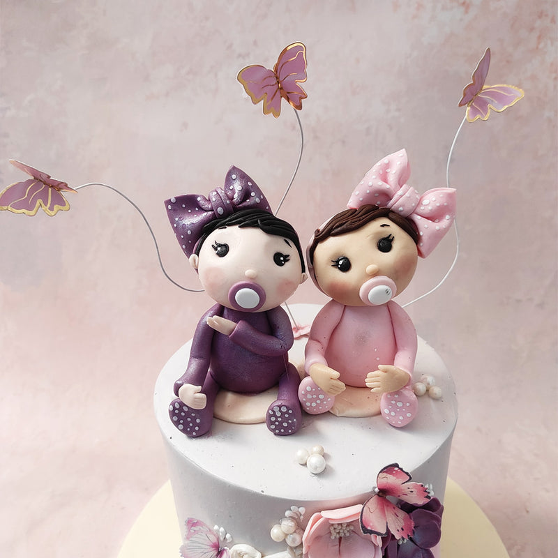 A figurine of both twins rests on top of this happy birthday twins cake, each one dressed in a vibrant coloured onesie with matching polka-dotted booties and bows! 