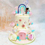 Atop the pastel-hued tiers of this Two Tier Fairy Garden Cake, a delightful rainbow arches behind a sweet fairy figure, with delicate wings and a flowing dress, stands beside toadstools. 