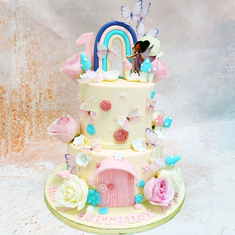 Atop the pastel-hued tiers of this Two Tier Fairy Garden Cake, a delightful rainbow arches behind a sweet fairy figure, with delicate wings and a flowing dress, stands beside toadstools. 