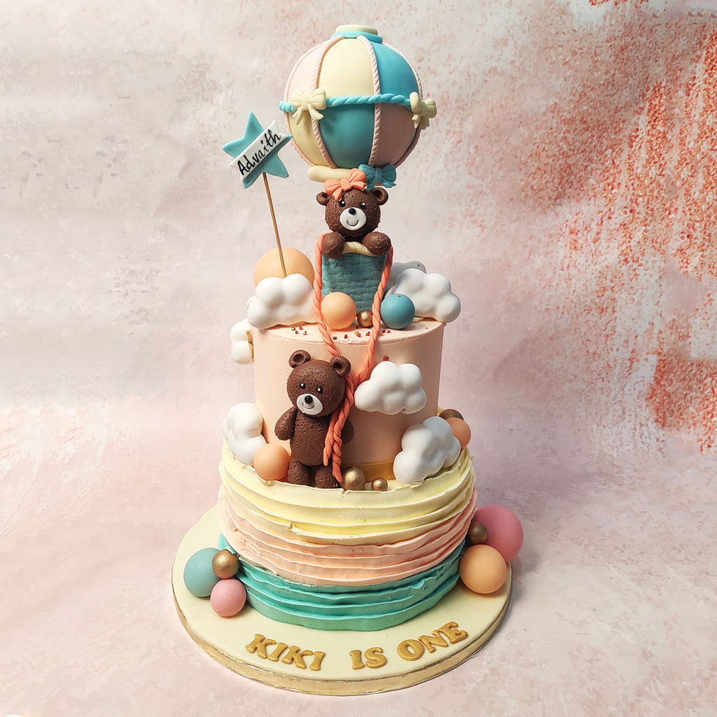 Embellished with chocolate spheres in shades of blue, pink, orange, and gold, this Two Tier Teddy Balloon Cake exudes playfulness and vibrancy.