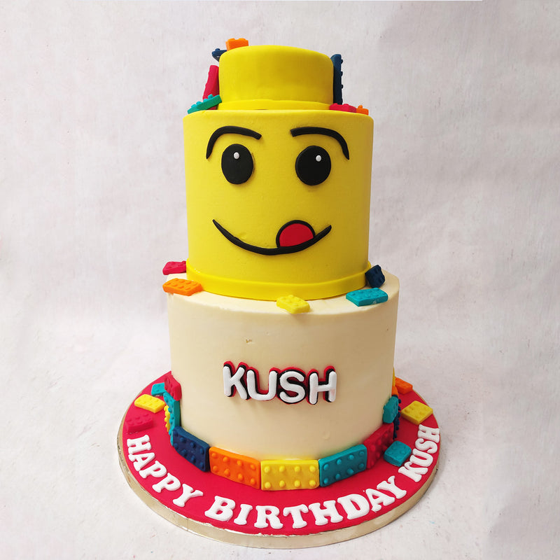 The top tier of this two tier Lego Theme cake is a giant Lego head with the emoji face sticking its tongue out, with a confetti rain of Lego blocks all over it.