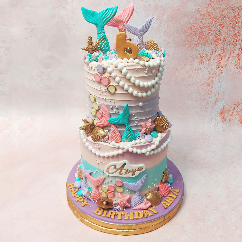 Both tiers of this Two Tier Mermaid Theme Cake boast a mesmerising spiral-textured pink buttercream base, reminiscent of the gentle currents that caress the ocean floor. 