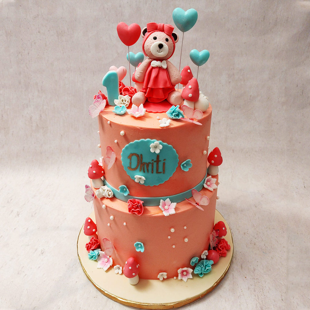 This garden theme cake features two pink bases, symbolising innocence and joy, adorned with an adorable teddy bear figurine on top. 