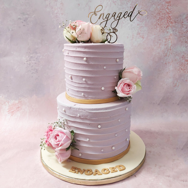 Each tier of this two tier purple engagement cake is delicately adorned in a light shade of mauve, exquisitely complemented by the presence of pearls and gold ribbons at the bottom. 