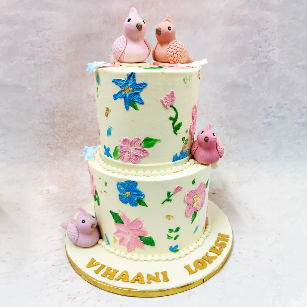 The design of this Two Tier Bird Theme Cake features a stunning hand-painted floral pattern, where delicate pink and vibrant blue blooms appear to dance across the ivory canvas.