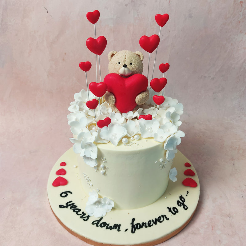 Atop the sweet symphony of this White Flowers Cake stands a beige teddy bear with a red heart clasped in its paws.