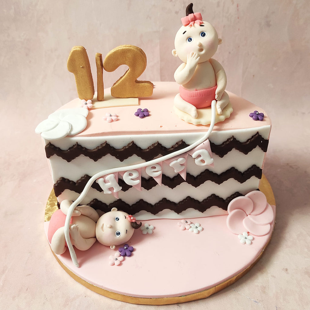 On top and at the bottom of this pink half birthday cake sits two charming figurines of a baby, each one holding one end of a banner flag. 