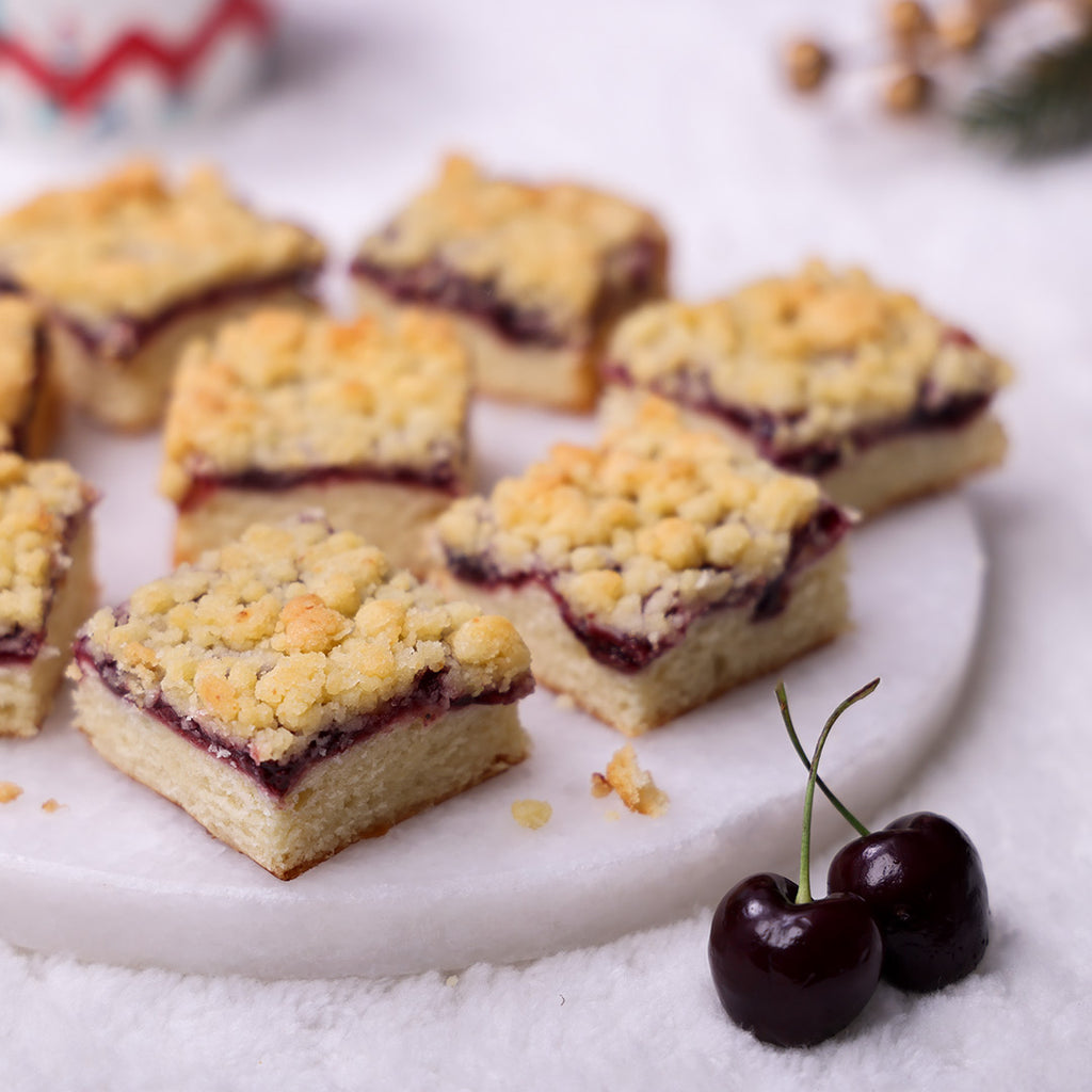 Assorted Cherry Crumble Cake squares on a marble plate, showing fruit filling and buttery crumb topping, garnished with fresh cherries.