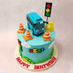 Two traffic lights can be spotted on this Tayo The Little Bus birthday cake for kids, one at the top next to a three-dimensional figurine of little Tayo himself who can be seen smiling in his parked spot near some orange traffic cones. 